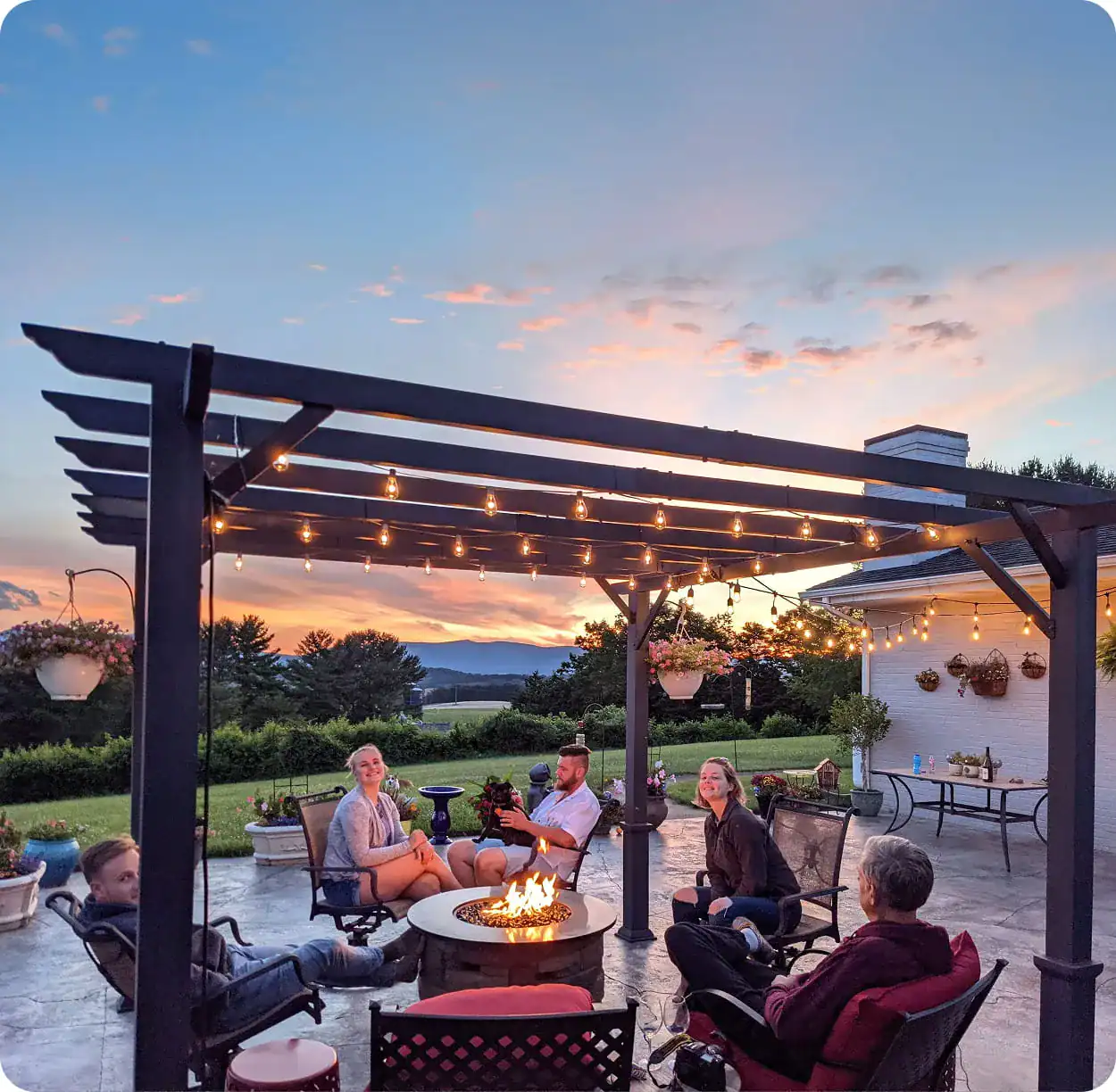backyard-pergola-fire-pit-sunset-gathering Family and friends relaxing around a fire pit under a pergola with string lights at sunset