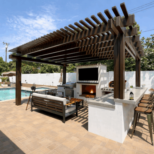 Poolside outdoor living area with pergola, outdoor kitchen, fireplace, bar seating, and lounge furniture.