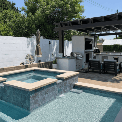 Modern backyard pool and spa with waterfall feature, outdoor kitchen, pergola, and lounge seating.