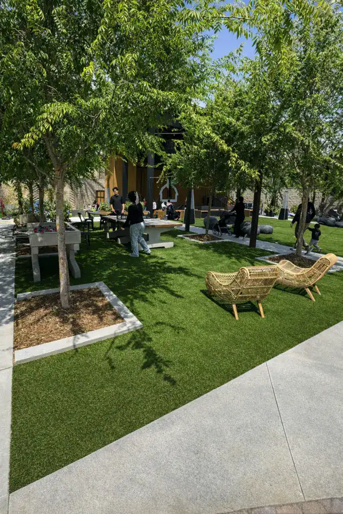 A community courtyard with mature trees, seating, and people playing ping-pong and foosball on a artificial turf surface.